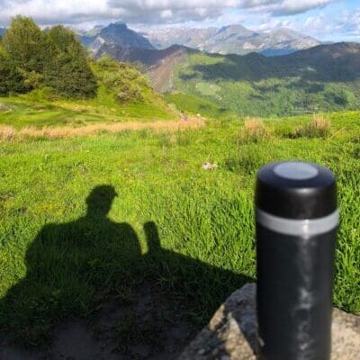 Paisaje montañoso con cielo azul y sombra de una persona sentada junto a un termo negro. Imagen que transmite pausa, reflexión y cuidado personal en el camino.