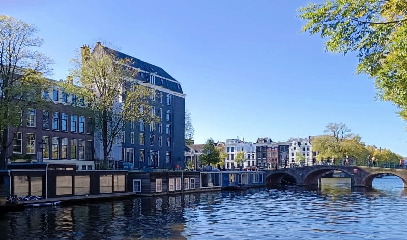 Vista de un canal en Ámsterdam con casas flotantes, edificios históricos y un puente peatonal bajo un cielo azul despejado.