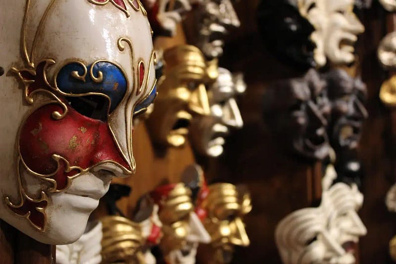 Máscaras venecianas tradicionales colgadas en una pared, con una en primer plano de colores blanco, rojo y azul, decorada con detalles dorados.