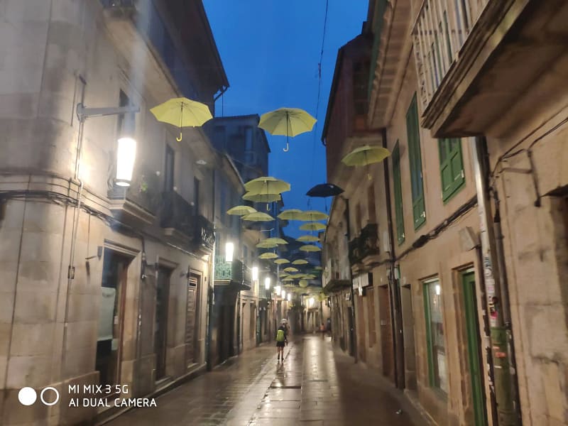 Calle estrecha y empedrada al anochecer, con paraguas amarillos colgando sobre el camino. Una persona camina al fondo bajo la iluminación de las farolas.